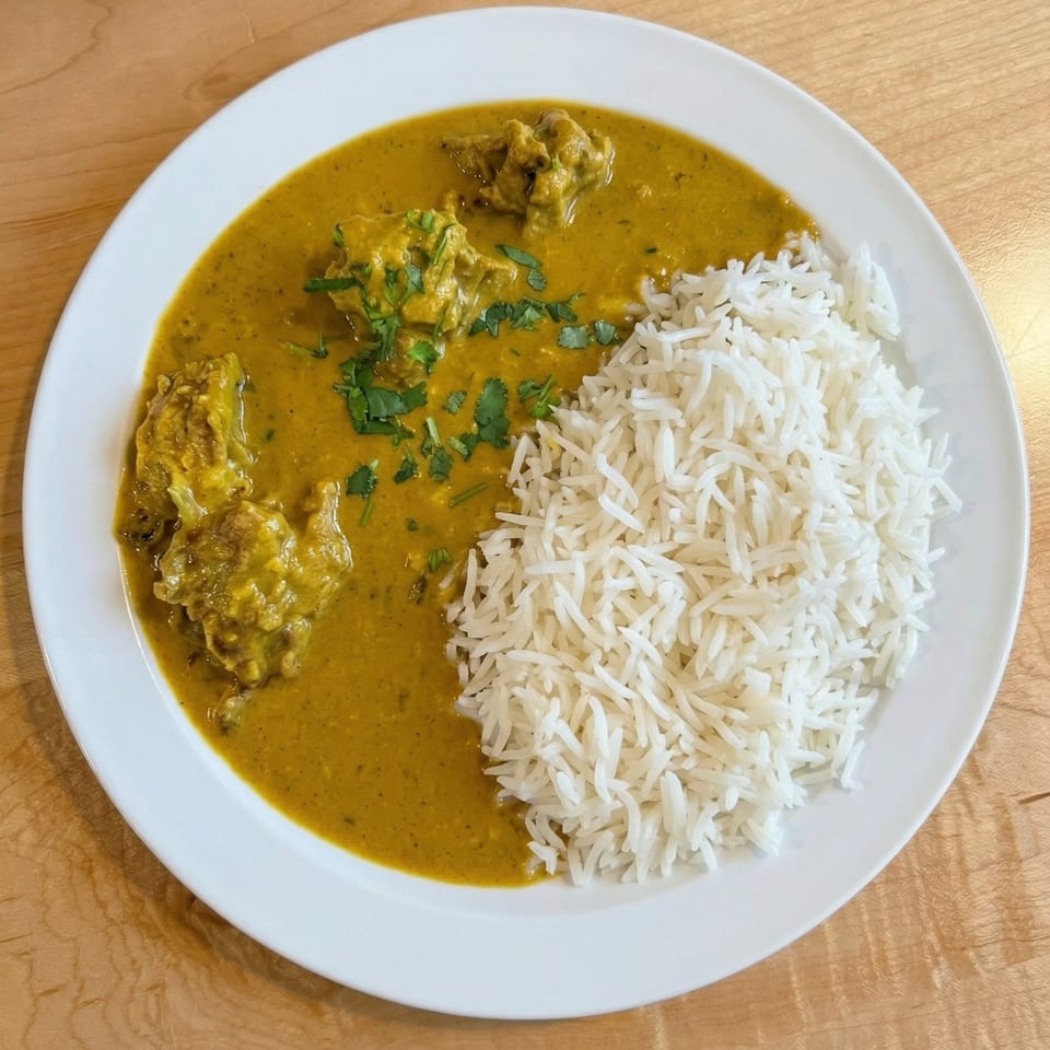 A white plate holds a serving of yellow curry with chicken and a side of white rice.