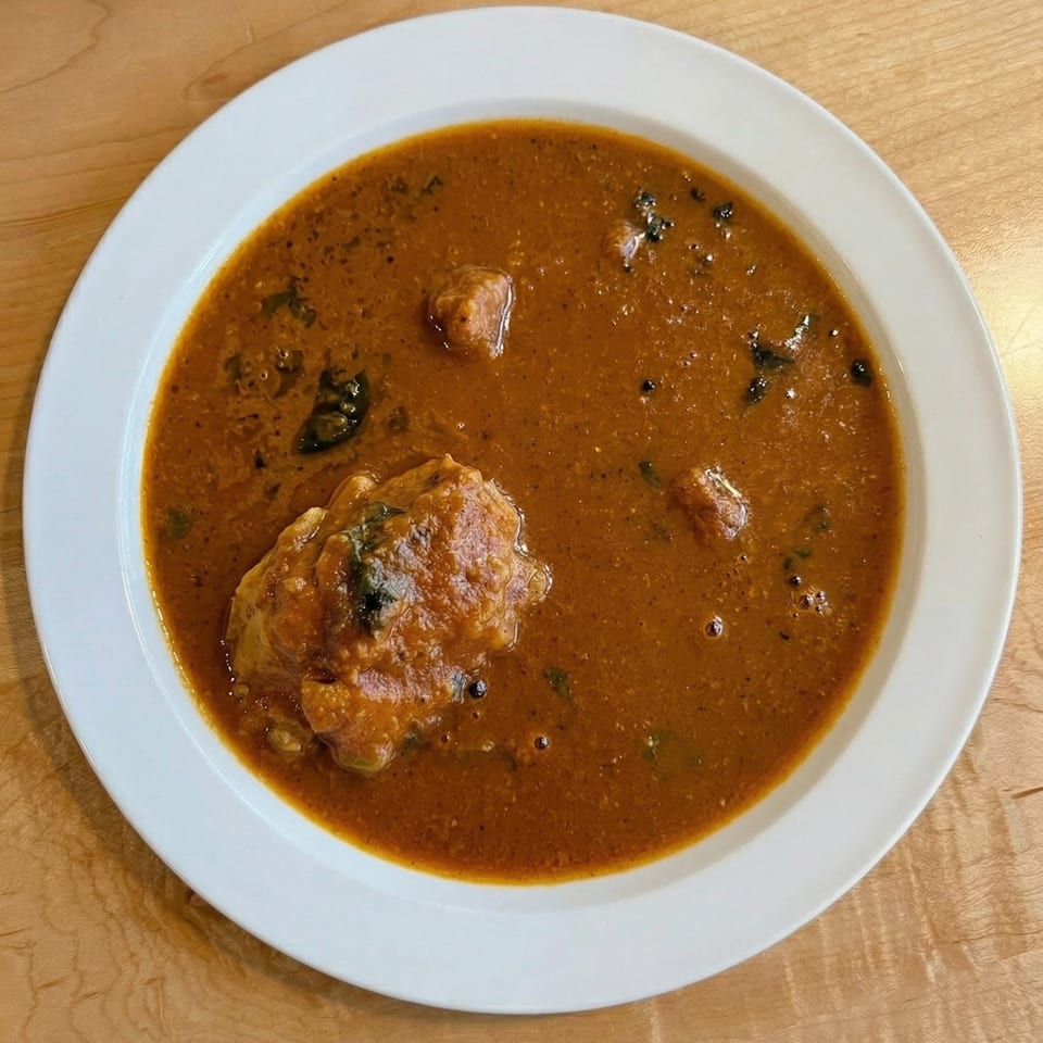 A white bowl filled with a rich, reddish-brown stew containing meat and dark leafy greens.